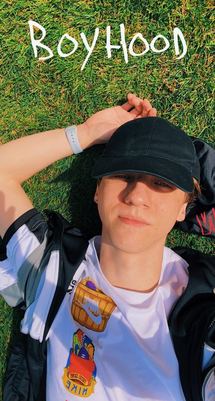 Me, lying down in the grass, with a black cap and a white shirt. My arm is behind my head. It's a sunny day.